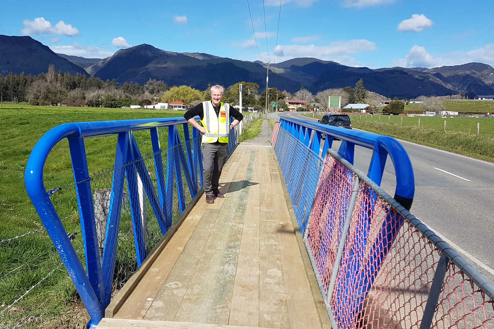 Man with yellow high-vis jacket standing on a cycle bridge with blue painted barriers on either side and a wooden planked surface.