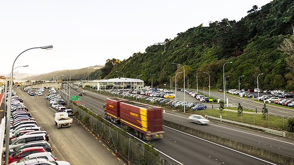 A large truck and cars travelling along a road, with cars parked in carparks either side of the road.
