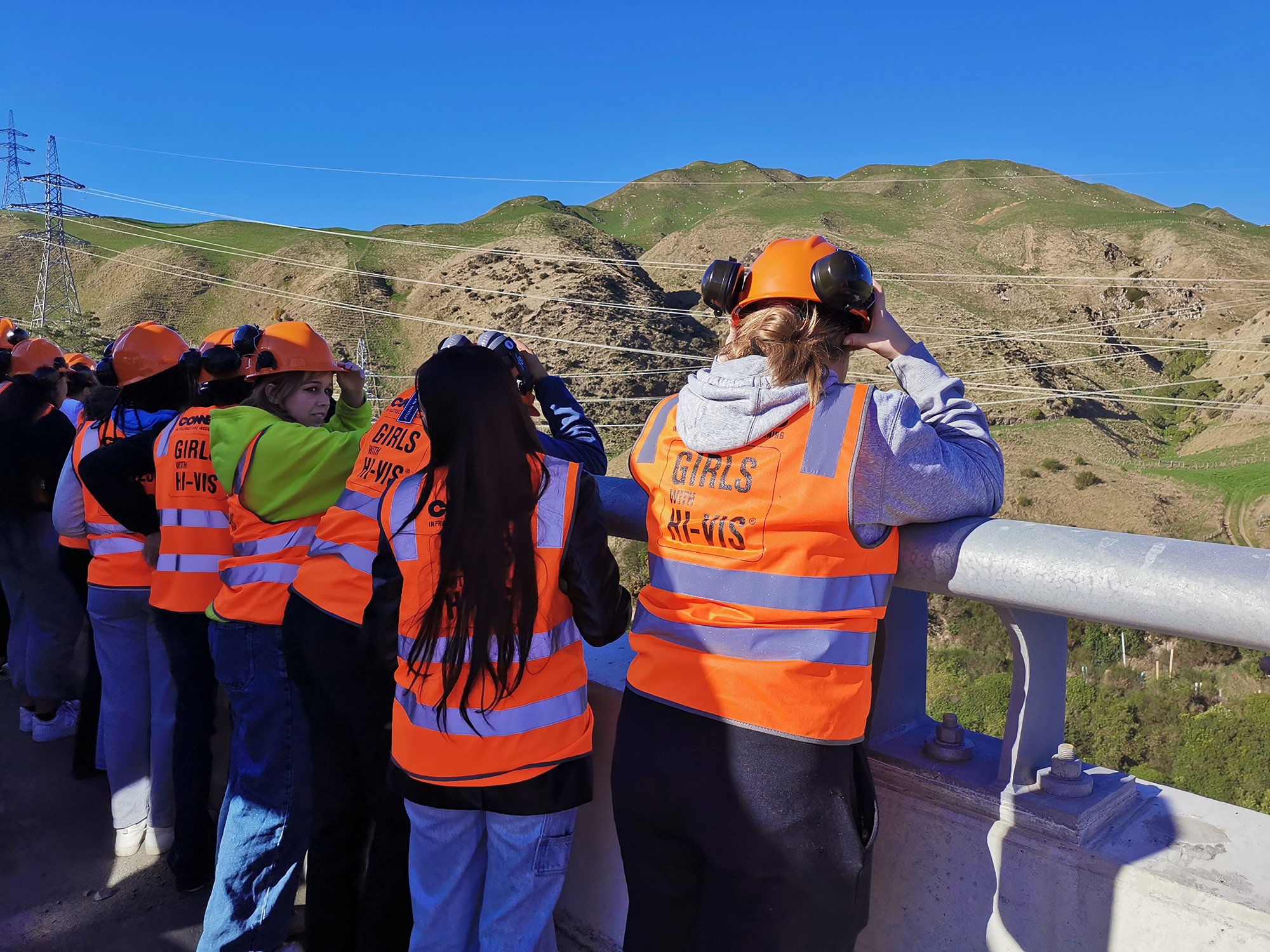 Girls with Hi Vis jackets and helmets standing on Te Ara a Toa bridge looking towards hills.