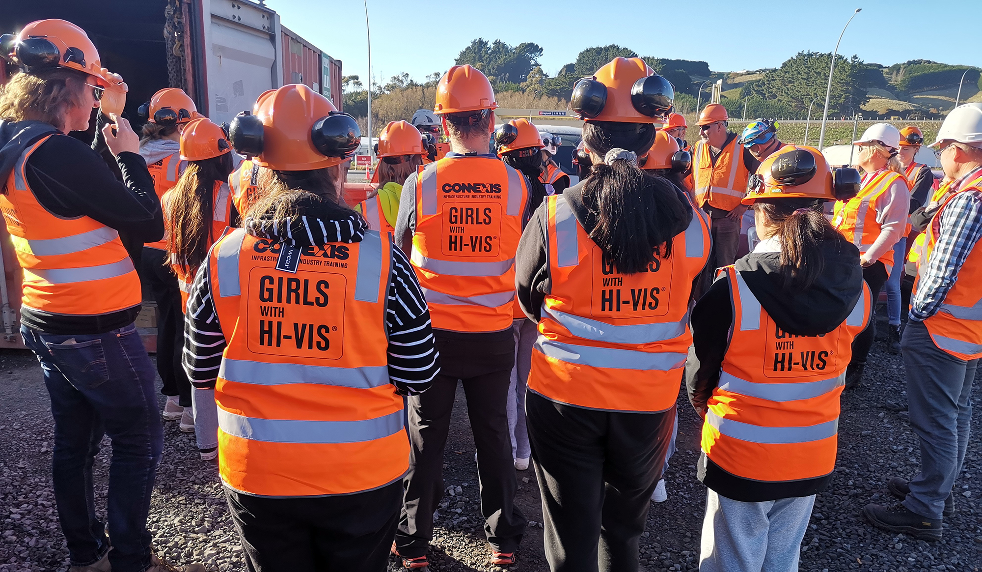 Girls wearing Hi Vis jackets, helmets and earmuffs standing with their backs to the camera.